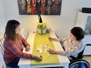 Eine Frau spielt mit einer anderen Frau im Rollstuhl an einem Tisch ein Brettspiel.