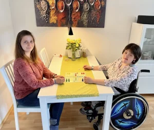 Eine Frau spielt mit einer anderen Frau im Rollstuhl an einem Tisch ein Brettspiel. Beide schauen in die Kamera.